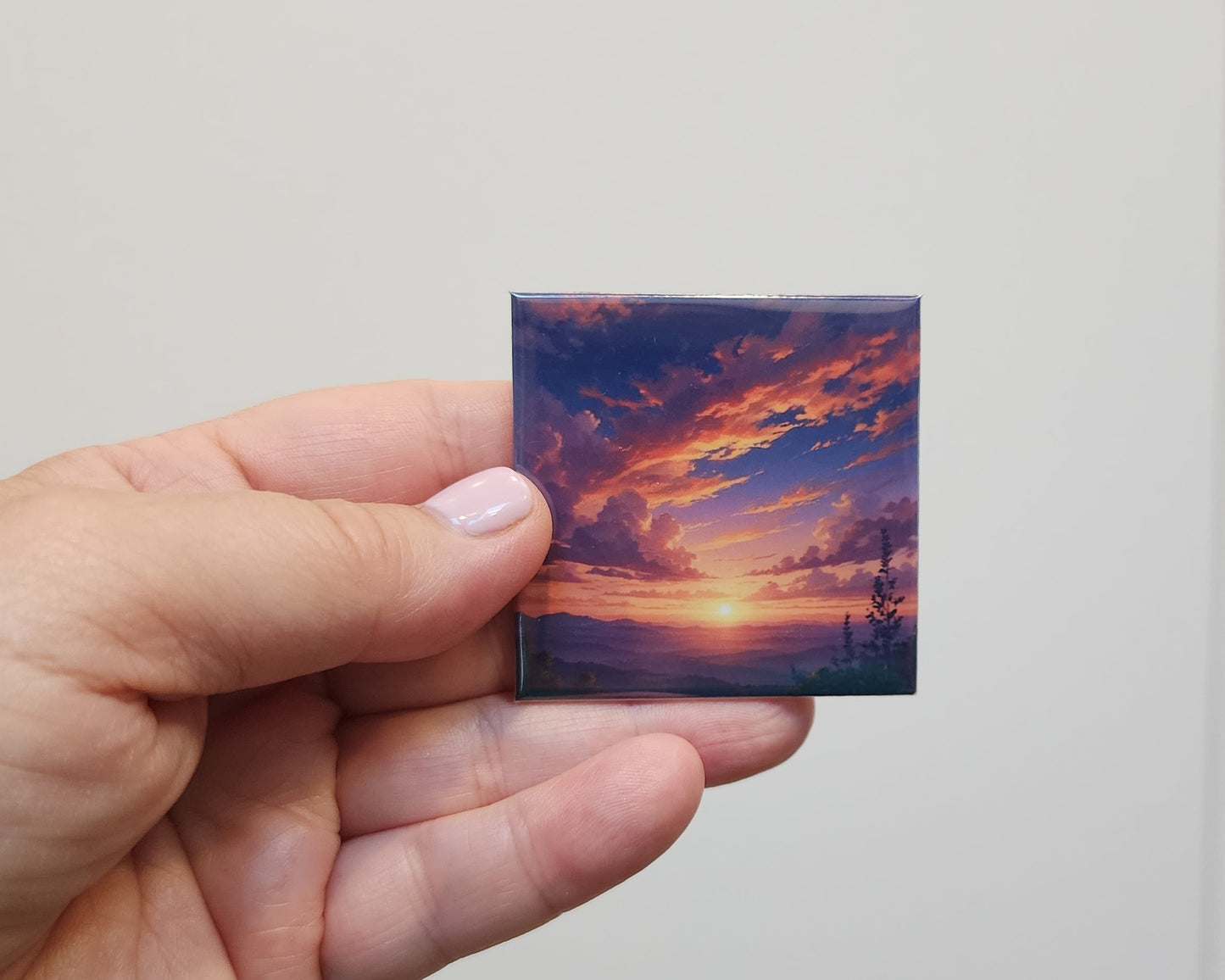 Hand holding a small square magnet of a sunset with palm trees against a plain background
